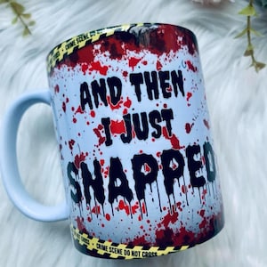 May include: A white ceramic mug featuring a red blood splatter pattern and yellow crime scene tape borders. The text "AND THEN I JUST SNAPPED" is printed in large, black, dripping letters across the centre of the mug, which has a light blue handle.