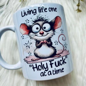 May include: White ceramic mug with a cartoon mouse illustration. The mouse has big eyes and red ears. The mug has the text "Living life one" and "Holy Fuck" at a time". The mouse is standing on a pink platform.