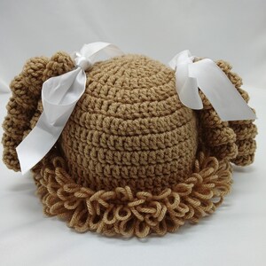 May include: A tan crochet baby hat with a curly fringe and two white satin bows. The hat is handmade and features a textured stitch pattern. The hat is designed to resemble a wig.