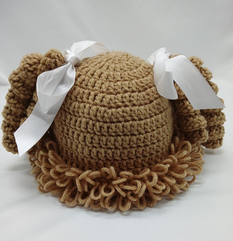 May include: A tan crochet baby hat with a curly fringe and two white satin bows. The hat is handmade and features a textured stitch pattern. The hat is designed to resemble a wig.