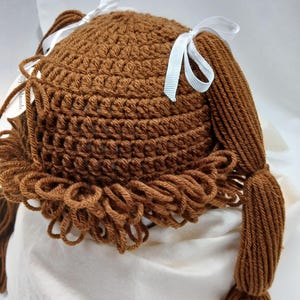 May include: A brown crocheted hat with two long braids and white ribbon bows. The hat has a textured pattern and a fringe of looped yarn around the bottom. The braids are made of the same brown yarn as the hat.
