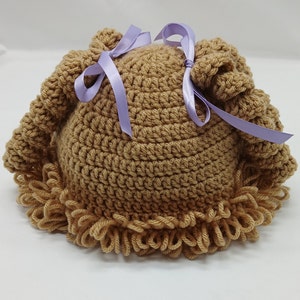 May include: A light brown, handmade crochet hat with a curly brim and two top knots. The hat is decorated with two lavender satin ribbons tied in bows. The hat is designed for babies and toddlers.