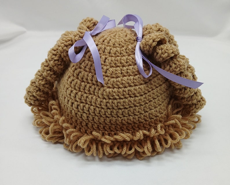 May include: A light brown, handmade crochet hat with a curly brim and two top knots. The hat is decorated with two lavender satin ribbons tied in bows. The hat is designed for babies and toddlers.