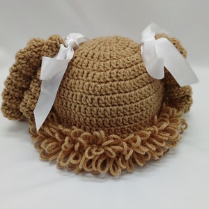 May include: A light brown crocheted hat designed to look like curly hair. The hat has a rounded crown and a ruffled edge, with white ribbon bows attached. This item is likely intended for a baby or child.