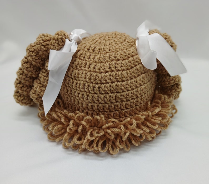 May include: A light brown crocheted hat designed to look like curly hair. The hat has a rounded crown and a ruffled edge, with white ribbon bows attached. This item is likely intended for a baby or child.