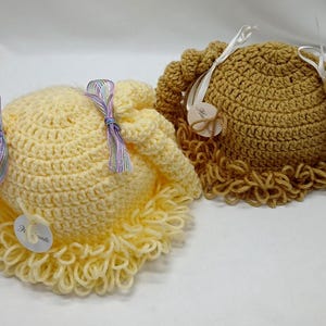 May include: Two crocheted hats are shown, one light yellow and one tan. They have a round shape with crocheted curls and are decorated with ribbons and small tags. The light yellow hat features lavender and blue ribbons.
