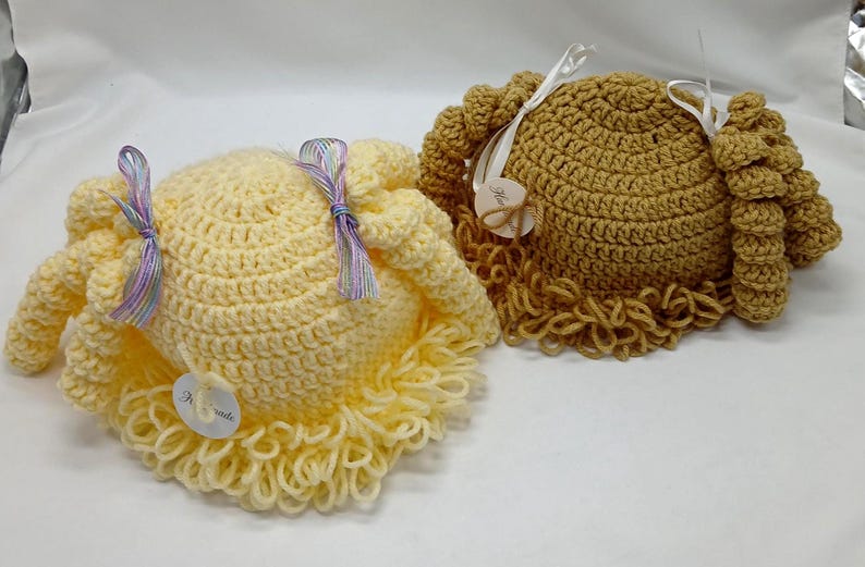 May include: Two crocheted hats are shown, one light yellow and one tan. They have a round shape with crocheted curls and are decorated with ribbons and small tags. The light yellow hat features lavender and blue ribbons.