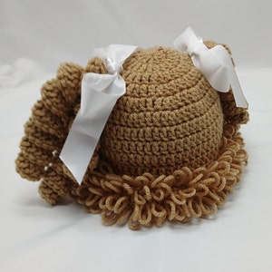 May include: A light brown crocheted baby hat, styled to look like a doll's hair, with a rounded crown and a fringed edge. Two white satin ribbons are tied on top. The hat is designed for infants.
