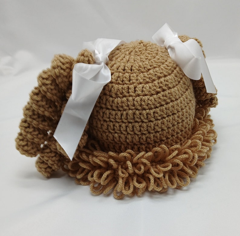 May include: A light brown crocheted baby hat, styled to look like a doll's hair, with a rounded crown and a fringed edge. Two white satin ribbons are tied on top. The hat is designed for infants.