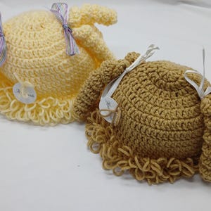 May include: Two handmade crochet hats, one light yellow and one tan, resembling doll wigs. Each hat has a curly fringe and is decorated with a ribbon and a small tag. The hats are displayed on a white surface.