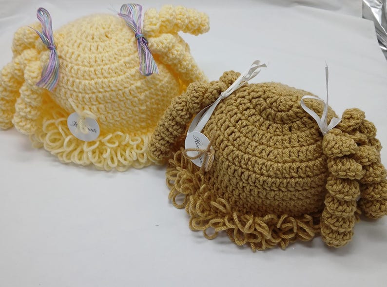 May include: Two handmade crochet hats, one light yellow and one tan, resembling doll wigs. Each hat has a curly fringe and is decorated with a ribbon and a small tag. The hats are displayed on a white surface.