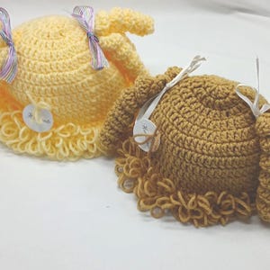 May include: Two handmade crochet hats. One is light yellow, the other is brown. Both have a round crown and a curly, fringed brim. Each hat has a ribbon and a tag attached. The hats are on a white surface.