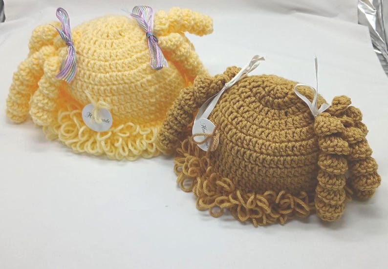 May include: Two handmade crochet hats. One is light yellow, the other is brown. Both have a round crown and a curly, fringed brim. Each hat has a ribbon and a tag attached. The hats are on a white surface.