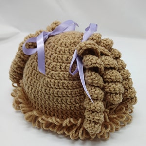 May include: A light brown crocheted baby hat with a curly fringe and two side accents tied with lavender ribbons. The handmade hat is designed for infants.