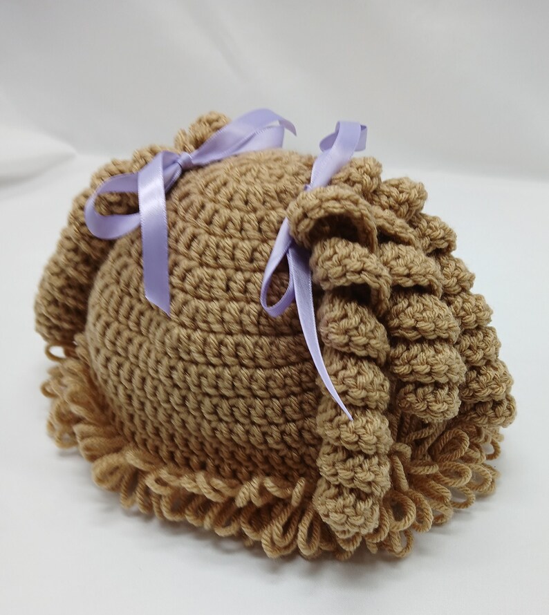 May include: A light brown crocheted baby hat with a curly fringe and two side accents tied with lavender ribbons. The handmade hat is designed for infants.