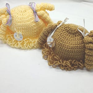 May include: Two handmade crochet hats in yellow and brown. Each hat features a round crown, a ruffled brim, and attached curly hair. The yellow hat has a lavender ribbon, and the brown hat has a white ribbon. Both hats have a small tag attached.