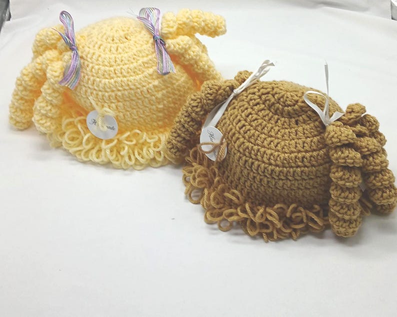 May include: Two handmade crochet hats in yellow and brown. Each hat features a round crown, a ruffled brim, and attached curly hair. The yellow hat has a lavender ribbon, and the brown hat has a white ribbon. Both hats have a small tag attached.