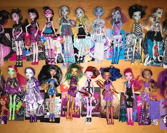 May include: A collection of Monster High dolls, each with unique hairstyles, outfits, and accessories. The dolls feature a variety of colors, including black, pink, blue, and green. Some dolls are holding accessories. The dolls are standing on a wooden surface.
