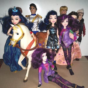 Puede incluir: Un grupo de muñecas de moda con cabello oscuro y atuendos coloridos. Una muñeca está montada en un caballo blanco. Las muñecas visten tonos de azul, morado, negro y rojo. Las muñecas están sobre una superficie marrón claro.