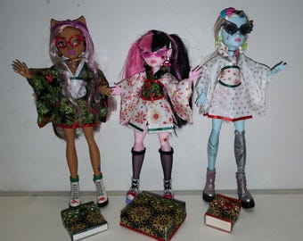 May include: Three fashion dolls wearing winter-themed outfits with a green, white and red colour scheme. The dolls are standing on small boxes with a gold and black pattern.