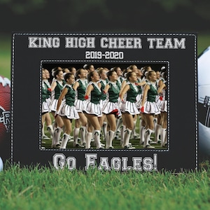May include: Black leather picture frame with white stitching. The frame has a photo of a cheerleading squad in green and white uniforms. The text "KING HIGH CHEER TEAM 2019-2020" and "GO EAGLES!" is printed on the frame.