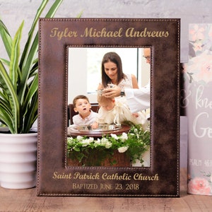 May include: A brown leather photo frame with a gold inscription that reads "Tyler Michael Andrews" and "Saint Patrick Catholic Church Baptized June 23, 2018". The frame contains a photo of a baby being baptized.