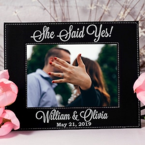 May include: Black leather picture frame with a white stitched border. The frame has a photo of a couple with the text "She Said Yes!" above the photo and "William & Olivia May 21, 2019" below the photo.