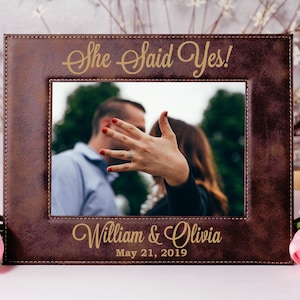 May include: A brown leather picture frame with a gold inscription that reads "She Said Yes!" and "William & Olivia May 21, 2019". The frame has a photo of a couple with the woman's hand in the foreground showing off her engagement ring.