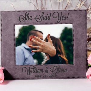 May include: Gray photo frame with a stitched border. The frame has a photo of a couple with the text "She Said Yes!" above the photo and "William & Olivia May 21, 2019" below the photo.