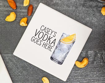 May include: A white square coaster with the text "CASEY'S VODKA GOES HERE" and a watercolor illustration of a cocktail with a lemon slice. Mixed nuts and a bowl of nuts are scattered around the coaster.