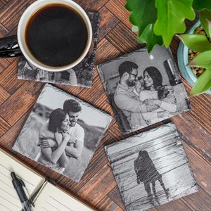 May include: Four black slate coasters with different black and white photographs printed on them. The coasters are arranged on a wooden surface with a cup of coffee and a potted plant.