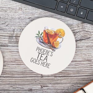 May include: A round, off-white coaster with the text "PHOEBE'S TEA GOES HERE". The coaster features a watercolor illustration of a cup of tea with lemon slices and a cinnamon stick. A keyboard and notebook are in the background.