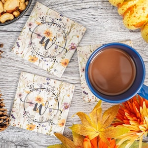 May include: Four floral coasters with a monogram in a wreath design. The coasters are on a wooden surface with a cup of coffee and fall-themed decorations.