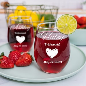 May include: Two wine glasses with red wine. The glasses are engraved with the names "Ashley" and "Haley" and the text "Bridesmaid" and "May 19, 2023".