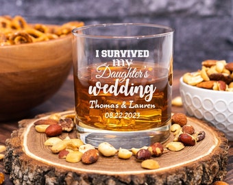 May include: A clear glass with the text "I survived my daughter's wedding Thomas & Lauren 08.22.2023" etched on the side. The glass is filled with a brown liquid and is sitting on a wooden surface with nuts and pretzels.