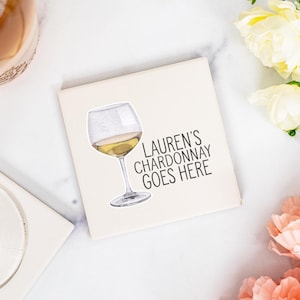 May include: A square, off-white coaster with a watercolor illustration of a wine glass filled with white wine. The text "LAUREN'S CHARDONNAY GOES HERE" is printed in black. The coaster is surrounded by flowers.