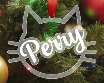 May include: Clear plastic Christmas ornament shaped like a cat's face with the name "Perry" written in white inside. The ornament is hanging from a red ribbon on a green Christmas tree.