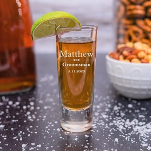 May include: A clear shot glass with the text "Matthew Groomman 5.11.2023" etched on the side. The glass is filled with a brown liquid and a lime wedge is resting on the rim.
