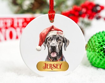 May include: A white ceramic Christmas ornament featuring a black dog wearing a red Santa hat with the year 2021 on it. The dog's name, "JERSEY", is written in gold on a gold banner.