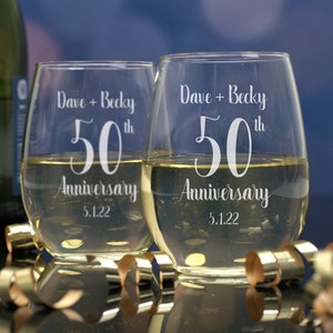 May include: Two clear glass wine glasses with the text "Dave + Becky 50th Anniversary 5.1.22" etched on them.