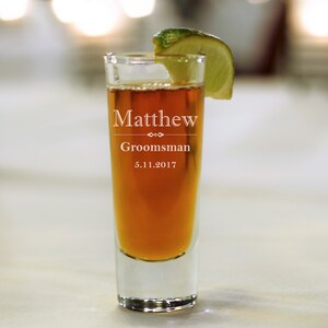 May include: A clear glass shot glass with a brown liquid inside. The glass is engraved with the text "Matthew Groomsman 5.11.2017". A lime wedge is on top of the glass.