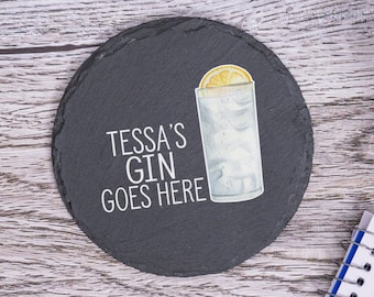 May include: A black slate coaster with the text "Tessa's Gin Goes Here" in white lettering. A watercolor illustration of a gin and tonic with a lemon wedge is on the right side of the coaster.