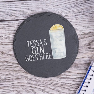May include: A black slate coaster with the text "Tessa's Gin Goes Here" in white lettering. A watercolor illustration of a gin and tonic with a lemon wedge is on the right side of the coaster.