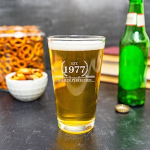 May include: A clear glass beer mug with a white inscription that reads "EST. 1977 Limited Reserve Aged to Perfection". The mug is filled with a golden-colored beer and is sitting on a dark gray surface.