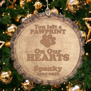 May include: A wooden Christmas ornament with the text "You left a pawprint on our hearts" and the name "Spanky" with the dates 2001-2012. The ornament is hanging on a Christmas tree.