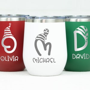 May include: Three stainless steel wine tumblers with personalized names and a Christmas-themed design. The tumblers are red, white, and green, and each features a different letter with a Santa hat. The names on the tumblers are Olivia, Michael, and David.