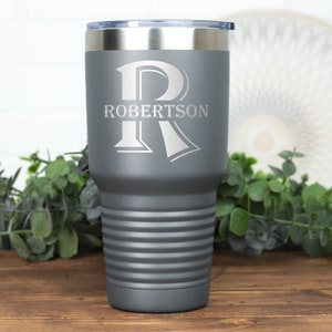 May include: A gray insulated tumbler with a silver rim and lid. The tumbler features the name "Robertson" and a large letter "R" etched in white. The tumbler has horizontal ridges and is set on a wooden surface.