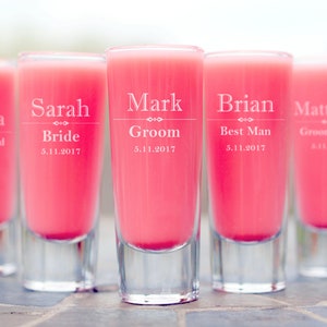 May include: Five clear glass shot glasses with pink liquid. Each glass is engraved with a name and title, such as "Olivia Bridesmaid" and "Mark Groom".