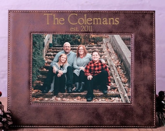 May include: A brown leather photo frame with a stitched border. The frame has a gold inscription that reads "The Colemans est. 2011". A family photo is displayed in the frame.