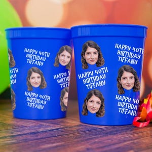 May include: Two blue plastic cups with the text "Happy 40th Birthday Tiffany" and a photo of a woman. A green balloon and colorful party streamers are in the background. The cups are on a wooden surface.